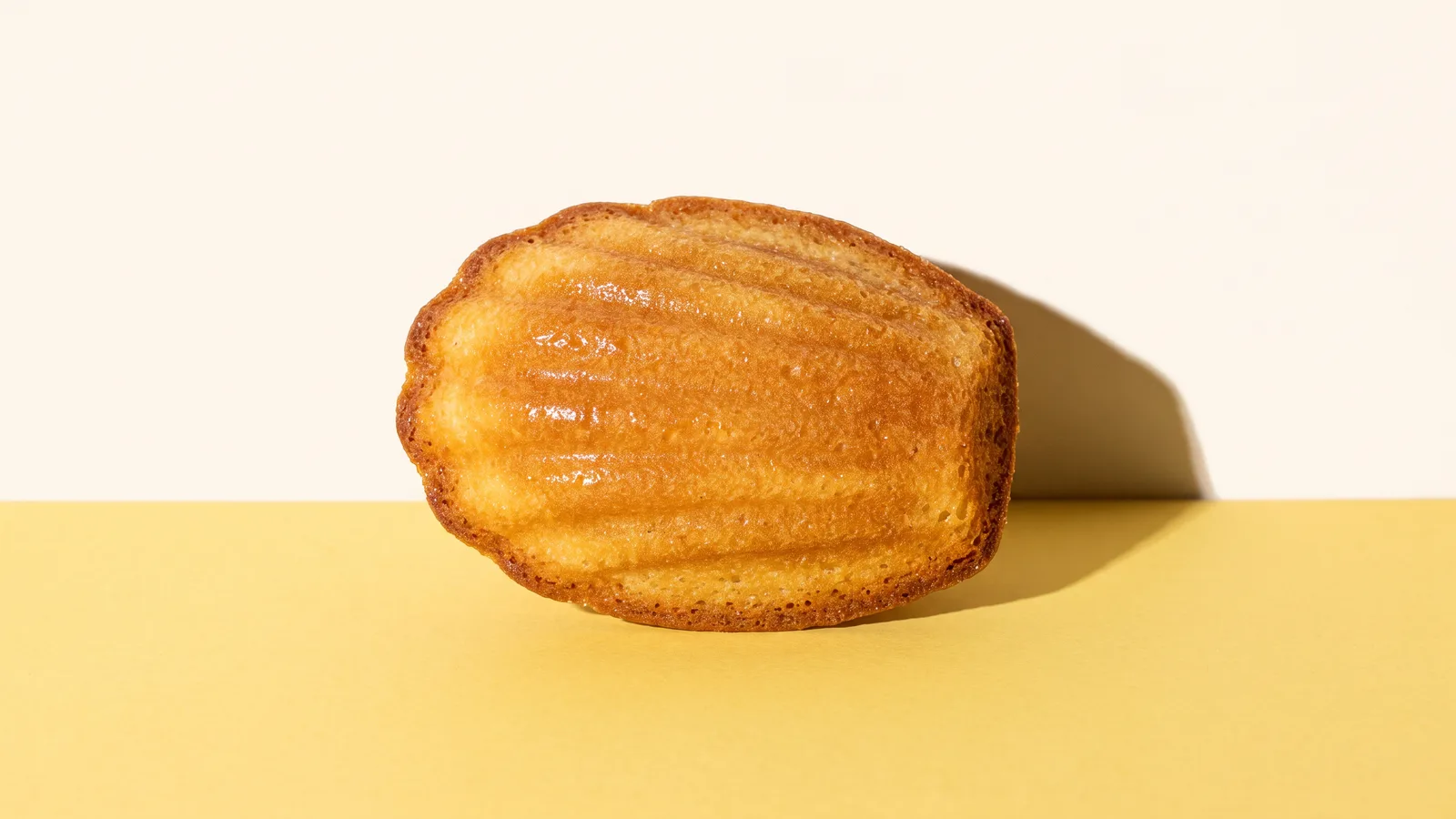 Close-up of a single madeleine showing its golden ridged shell side and darker crisp edges on a butter yellow surface