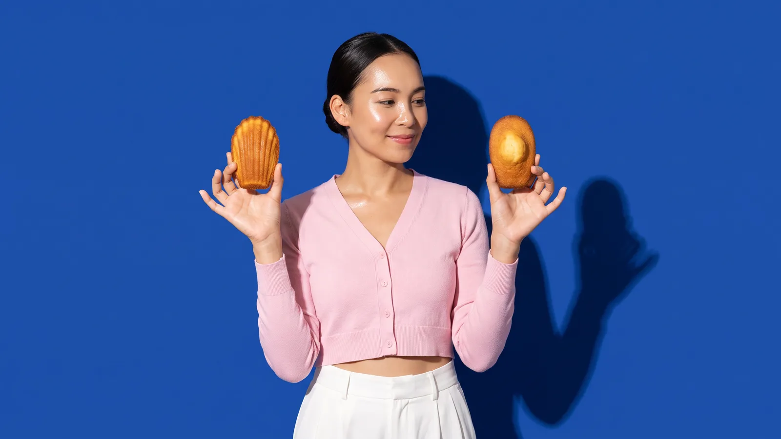 A smiling Thai woman in a pink cropped cardigan holds up a madeleine in each hand, comparing them, against a cobalt blue wall