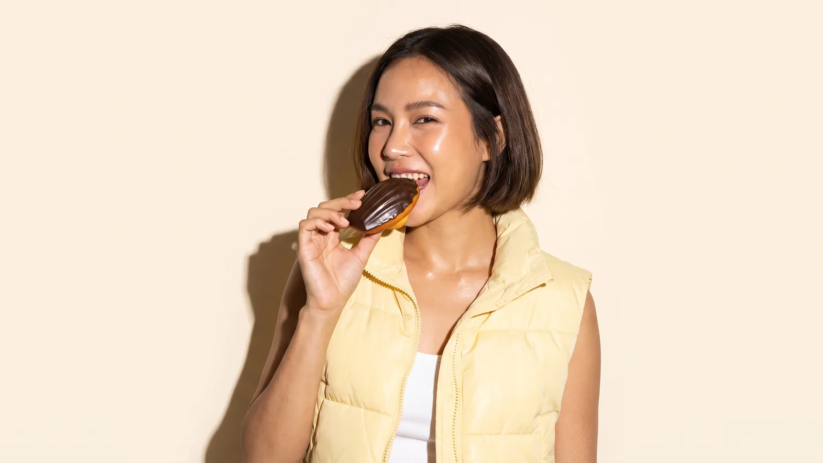 A Thai woman with a bob in a butter yellow puffer vest bites into a dark-coated madeleine against a cream wall