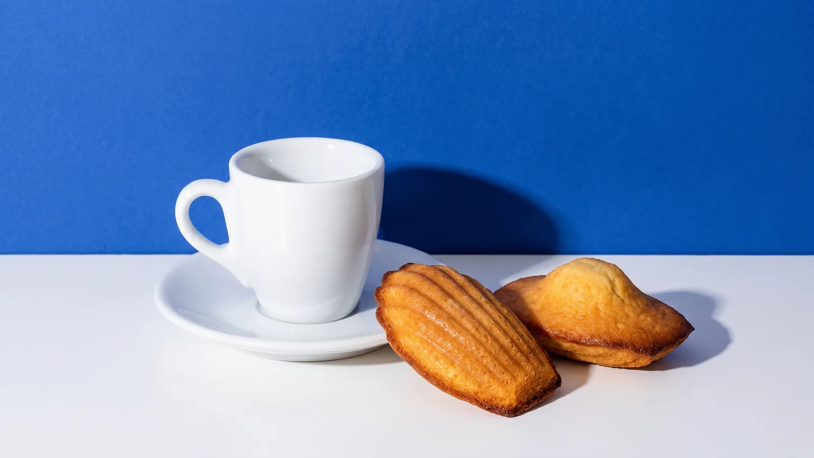 A plain white cup and saucer with two madeleines beside it on a white table against a cobalt blue wall