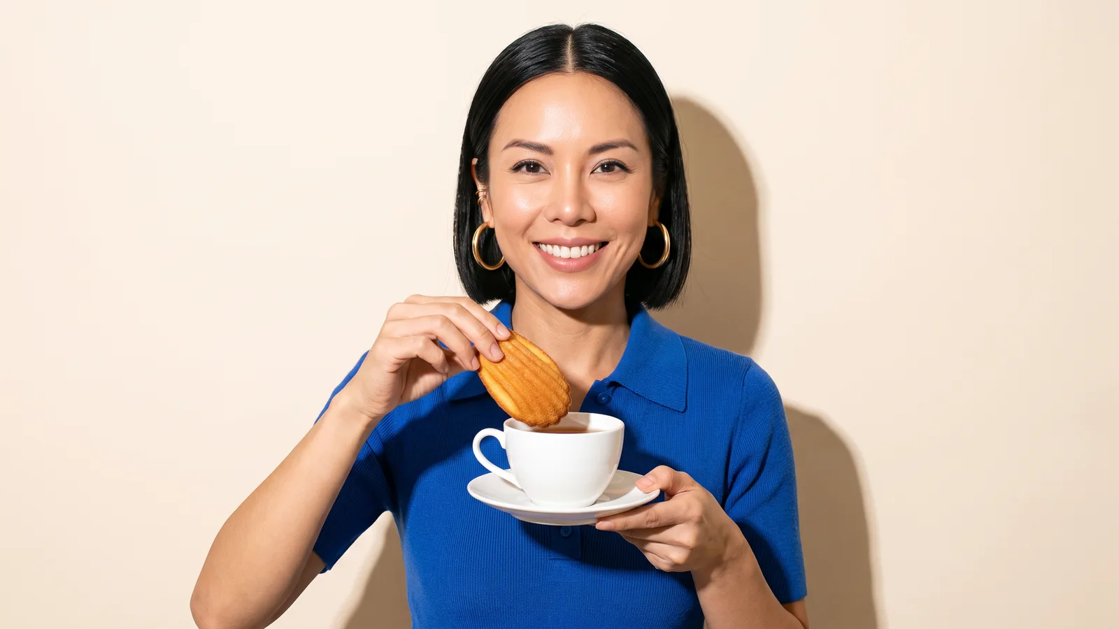 A Thai woman in a cobalt knitted polo dips a madeleine into a plain white teacup against a cream wall