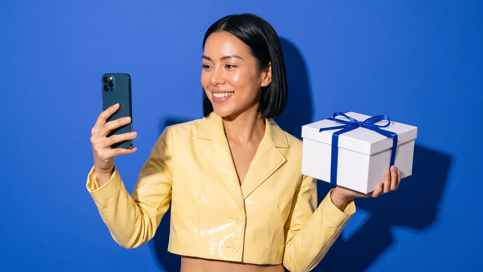 A Thai woman in a butter yellow cropped jacket smiles at her phone while holding a closed white gift box tied with a cobalt ribbon against a cobalt blue wall