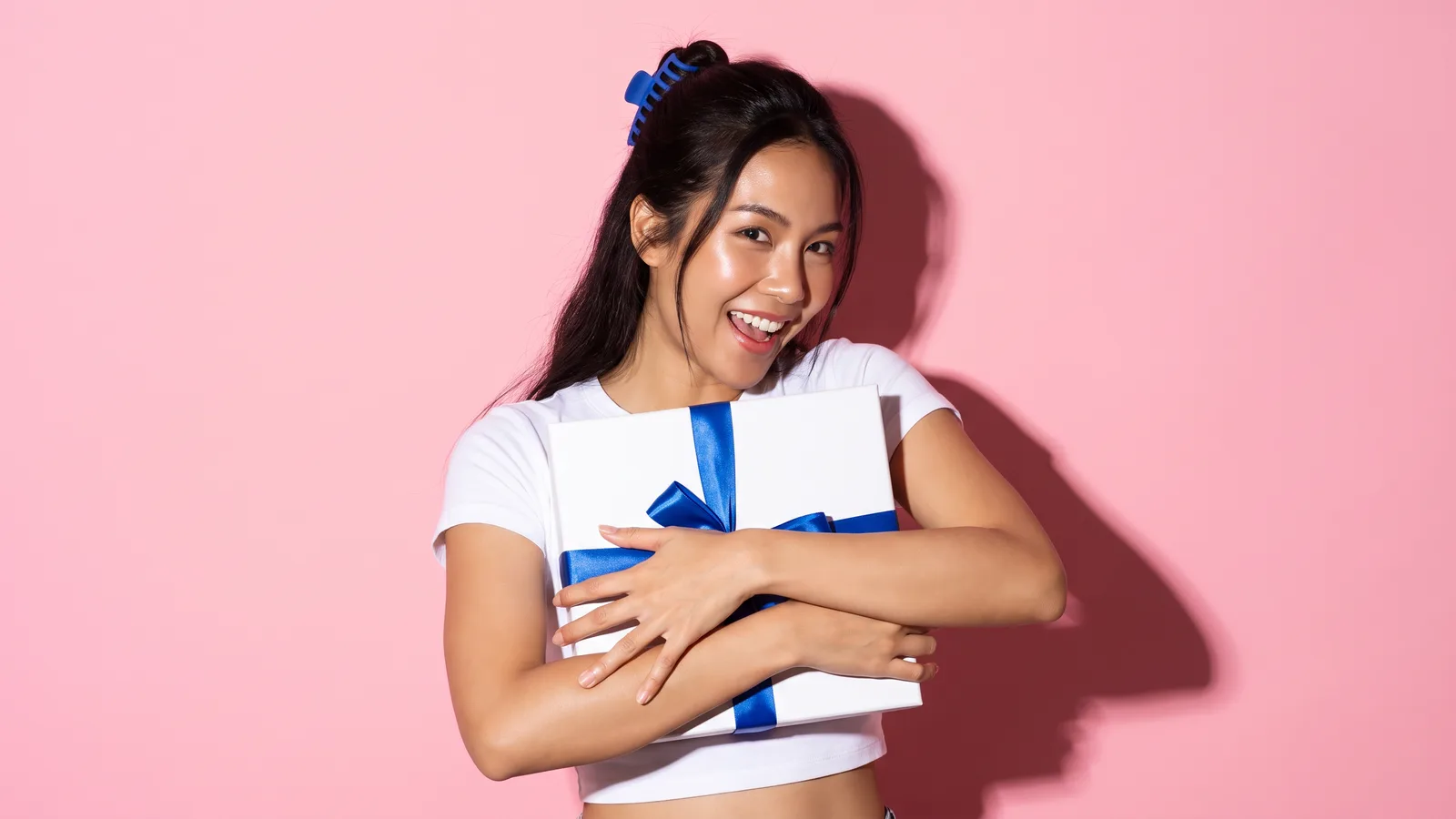 A Thai woman with a cobalt hair clip in a white cropped tee hugs a closed white gift box with a cobalt ribbon, smiling in surprise, against a pink wall
