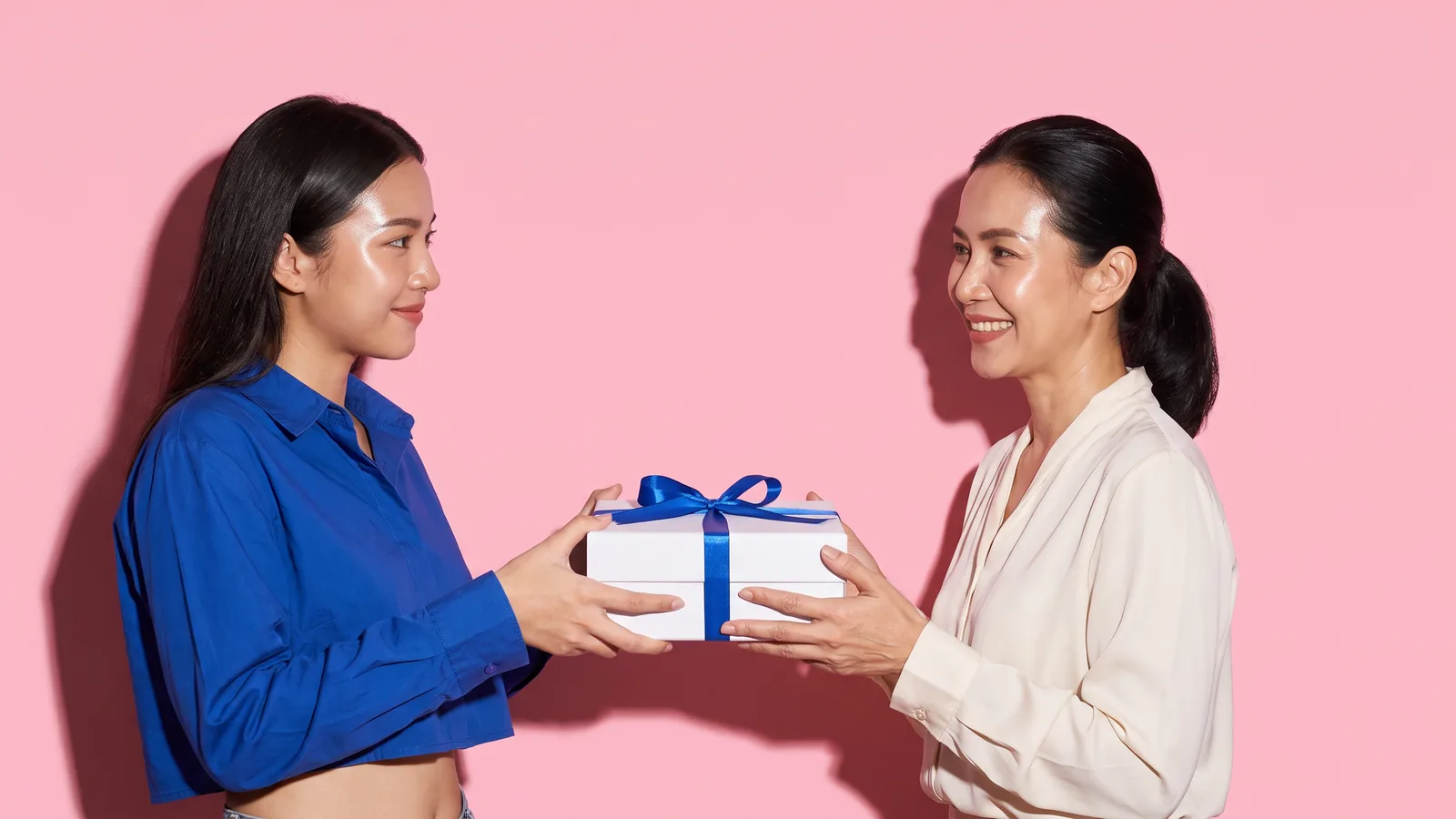 A young Thai woman in a cobalt shirt hands a white gift box with a cobalt ribbon to an older Thai woman in a cream blouse, both using both hands, against a pink wall