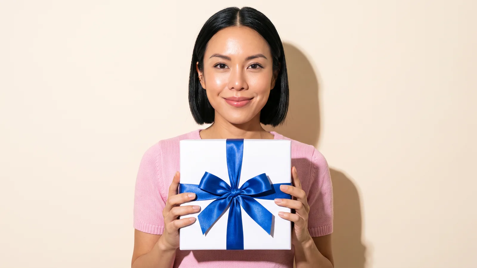 A smiling Thai woman in a pink knit top holds a white box tied with a cobalt satin bow in front of her against a cream wall