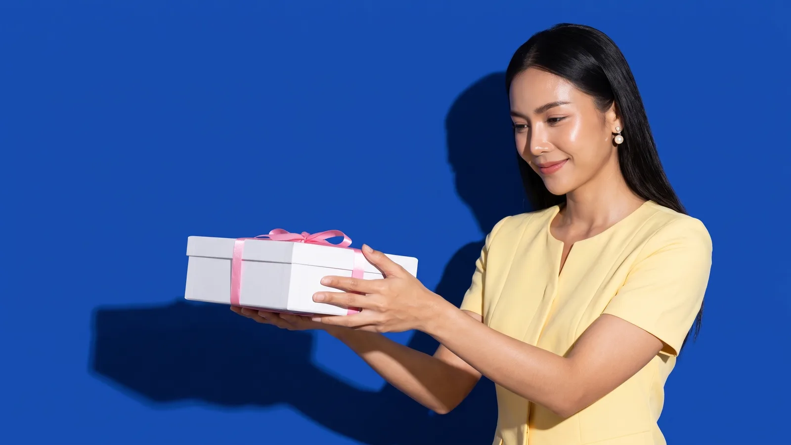 A Thai woman in a butter yellow top holds out a white gift box tied with a pink ribbon with both hands against a cobalt blue wall