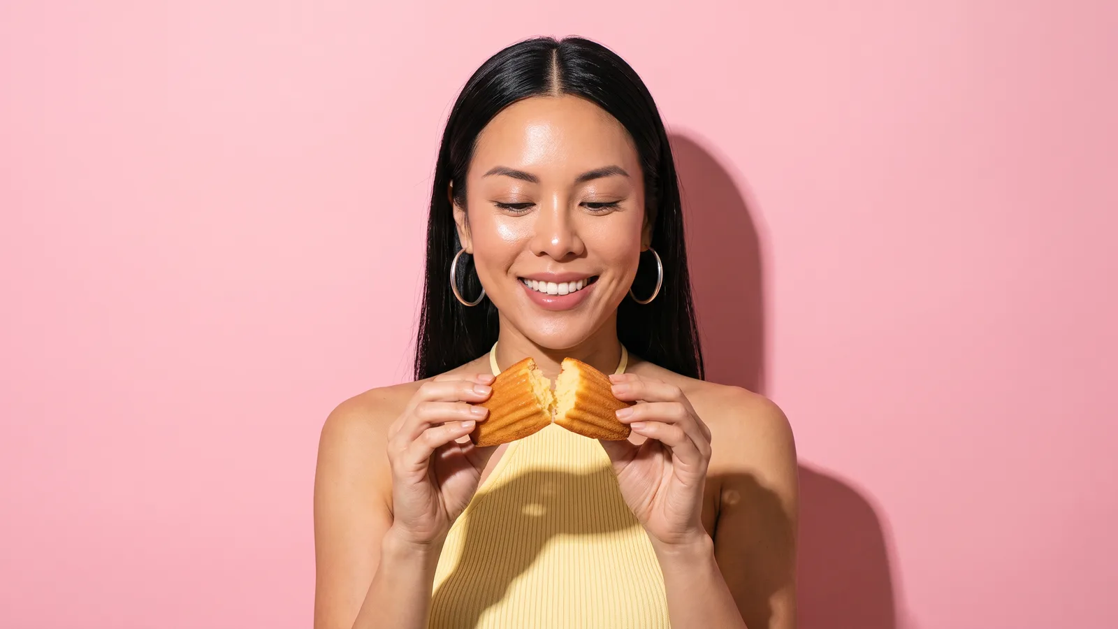 A smiling Thai woman with gold hoops breaks a madeleine in half with both hands against a pink wall