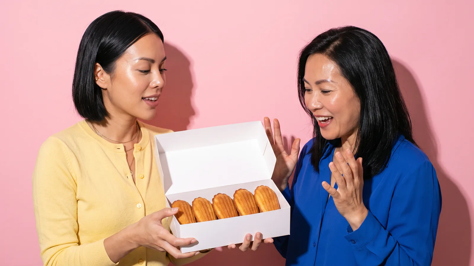 A young Thai woman in a butter yellow cardigan shows an open white box of five madeleines to a delighted older Thai woman in a cobalt blouse against a pink wall