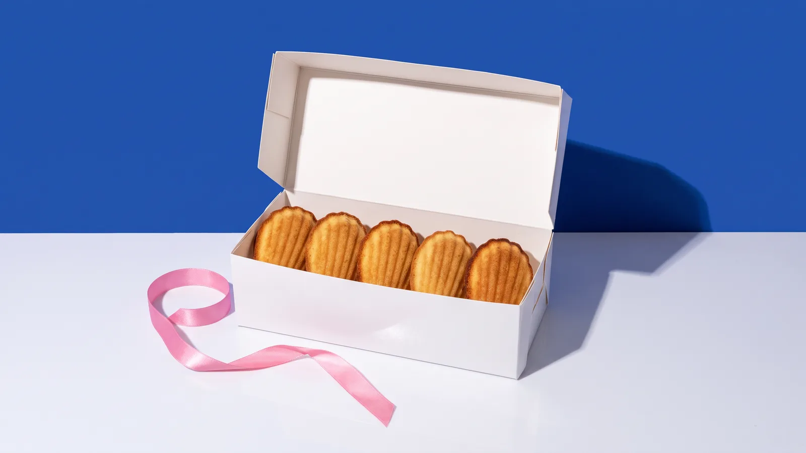 An open white box of five madeleines on a white surface beside a loose pink ribbon against a cobalt blue wall