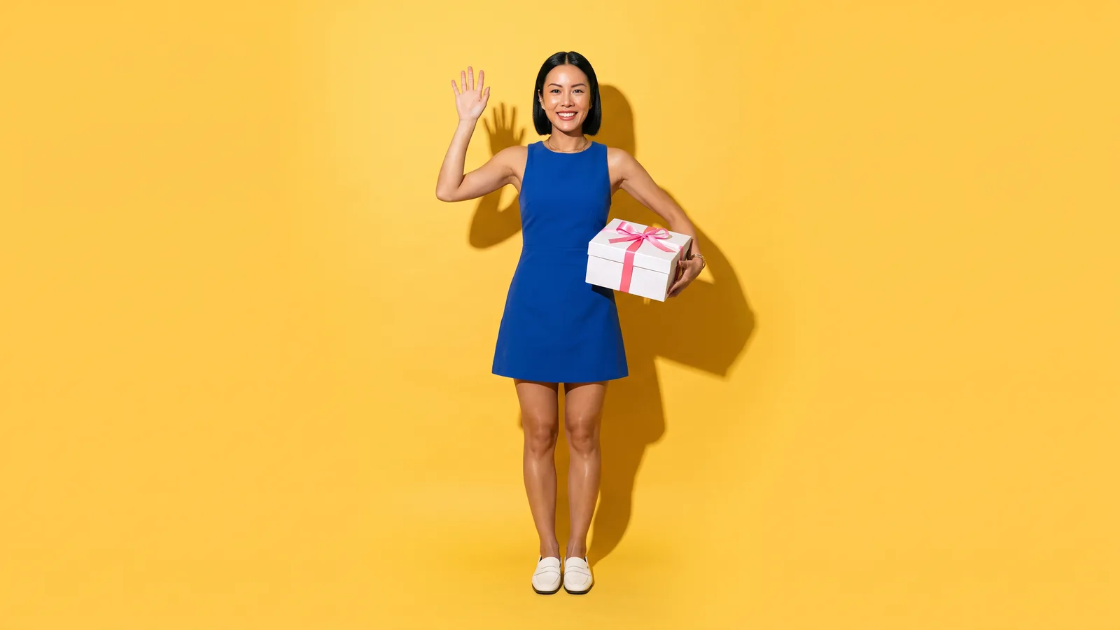 A smiling Thai woman in a cobalt mini dress and white loafers waves while carrying a white gift box with a pink ribbon against a butter yellow wall