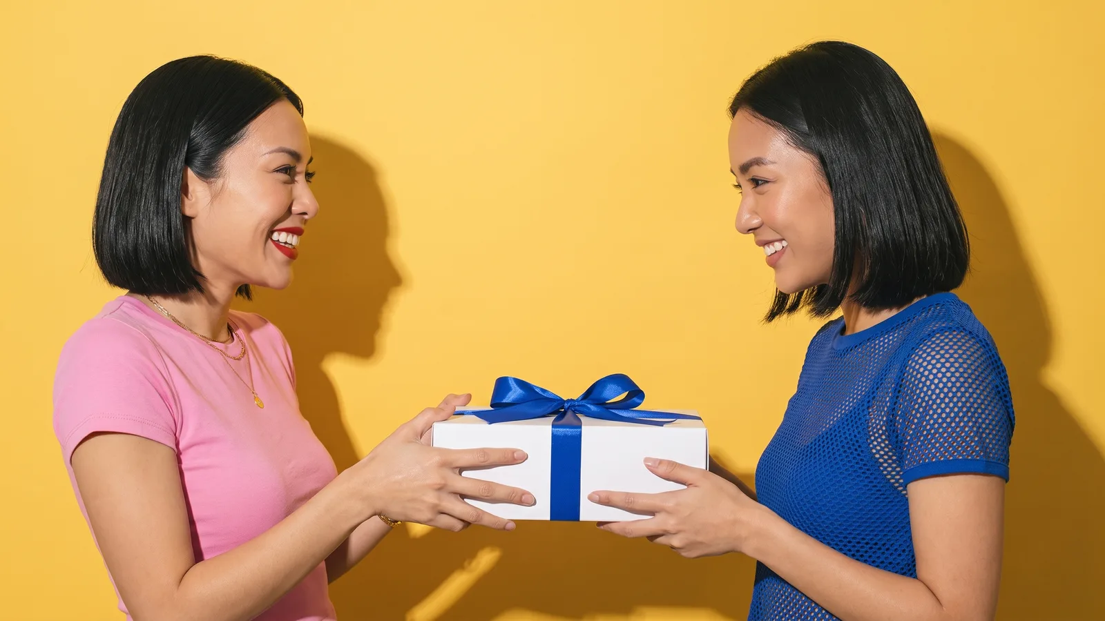 A Thai woman in a pink tee hands a white gift box tied with a cobalt ribbon to a friend in a cobalt mesh top against a butter yellow wall