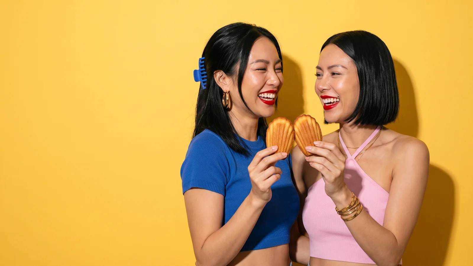 Two laughing young Thai women, one in a cobalt cropped knit with a cobalt claw clip, the other in a pink halter top with gold bangles, touch two golden madeleines together like a toast against a butter-yellow wall