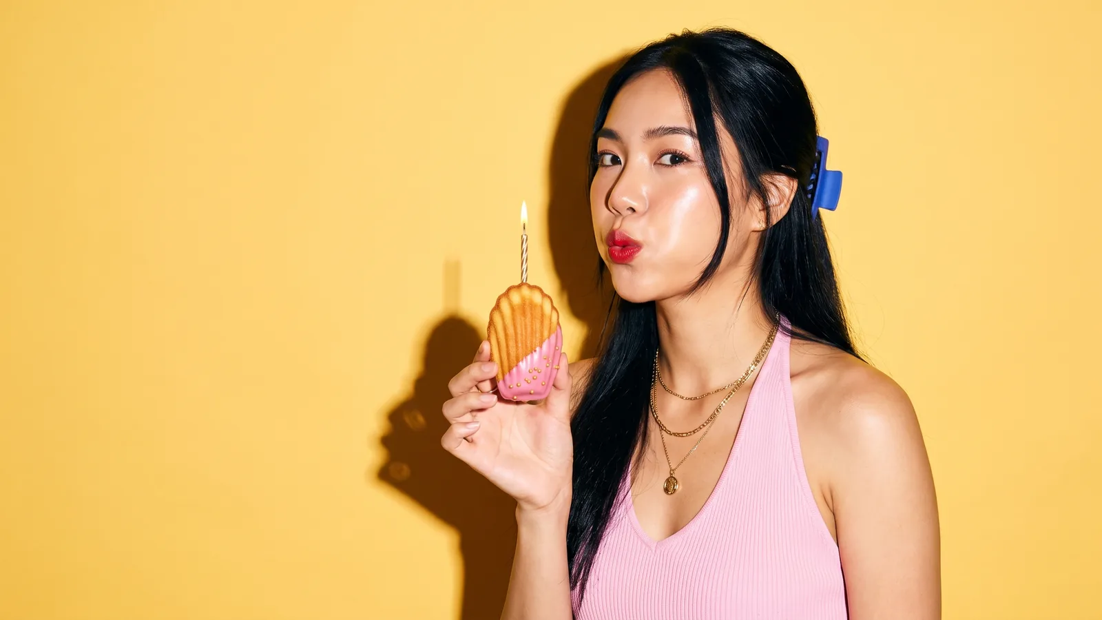 A young Thai woman in a pink halter top and a cobalt hair clip purses her lips to blow out a single candle on a madeleine half-dipped in pink glaze with gold sprinkles, against a butter-yellow wall