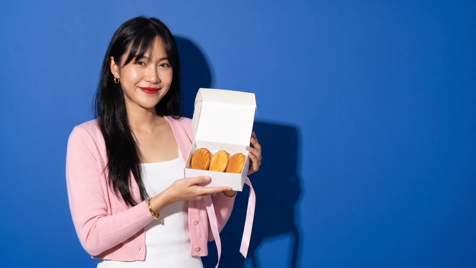 A smiling young Thai woman with long black hair, red lips and gold hoops, in a pastel pink cardigan, holds an open plain white box of three golden madeleines with a pale pink ribbon against a cobalt blue wall