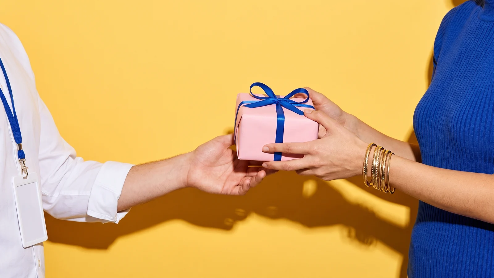 Two hands pass a small gift wrapped in pink paper with a cobalt blue ribbon against a bright yellow wall, one person in a white office shirt with a blank lanyard card, the other in a cobalt knit with gold bangles