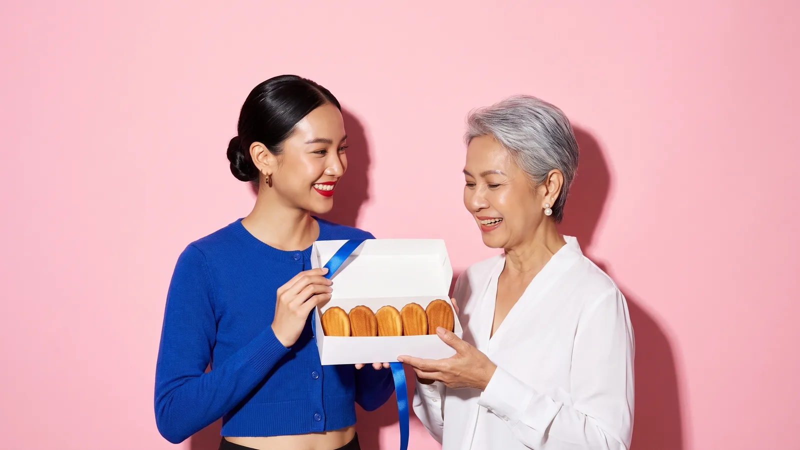 A smiling young Thai woman in a cobalt cardigan offers an open plain white box of five golden madeleines with a cobalt ribbon to a delighted older Thai woman with silver hair, against a pink wall