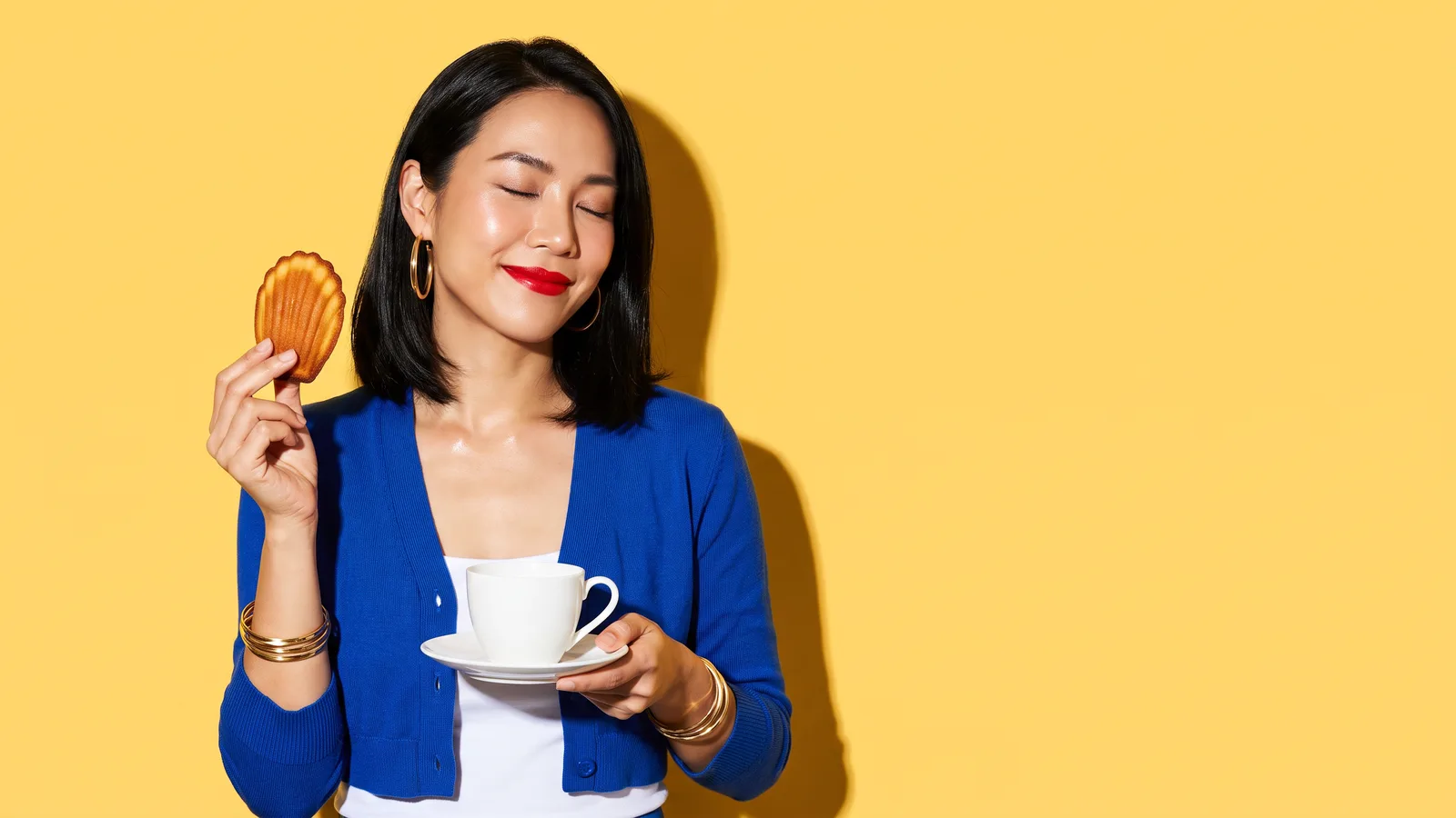 A Thai woman with a black bob and red lips, in a cobalt blue cardigan and gold bangles, closes her eyes and smiles as she holds up a golden madeleine in one hand and a white teacup on a saucer in the other, against a sunny yellow wall